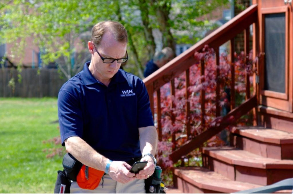 A WIN home inspector franchise owner looks down at his mobile phone while inspecting a backyard.