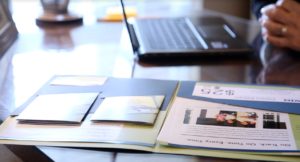 A folder of marketing materials sits on the desk of a real estate agent, with an out-of-focus laptop computer and hand in the background.