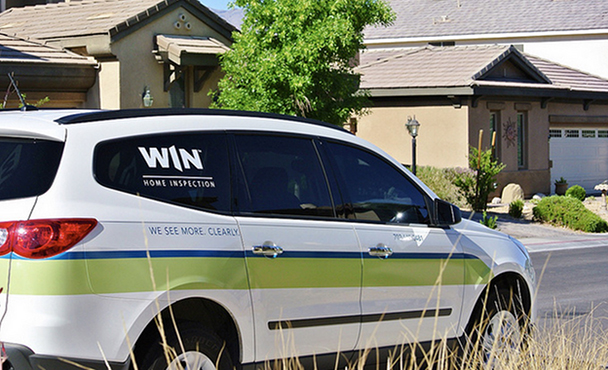 A WIN Home Inspection minivan with the “We see more. Clearly.” tagline is parked next to a patch of tall grass in front of a row of homes.
