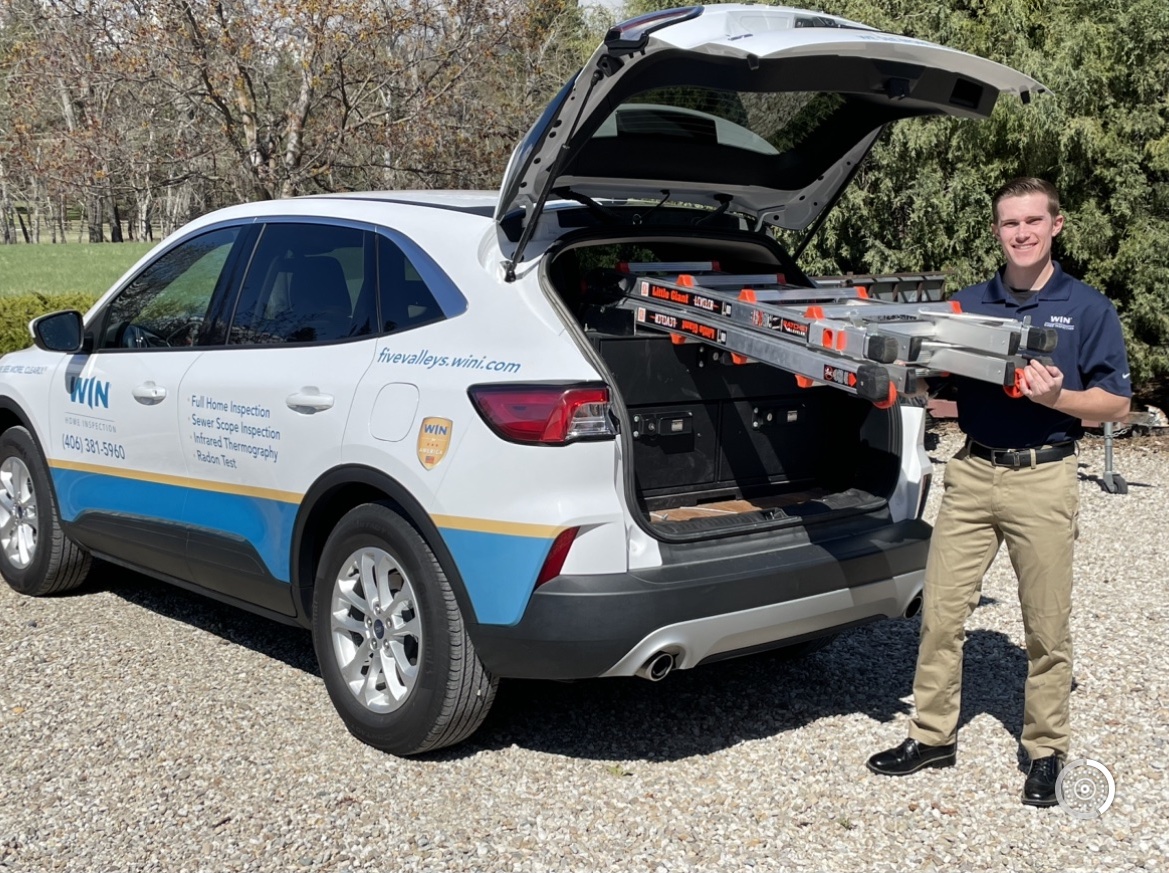 A home inspector stands next to an SUV with the trunk open, holding an extendable ladder.