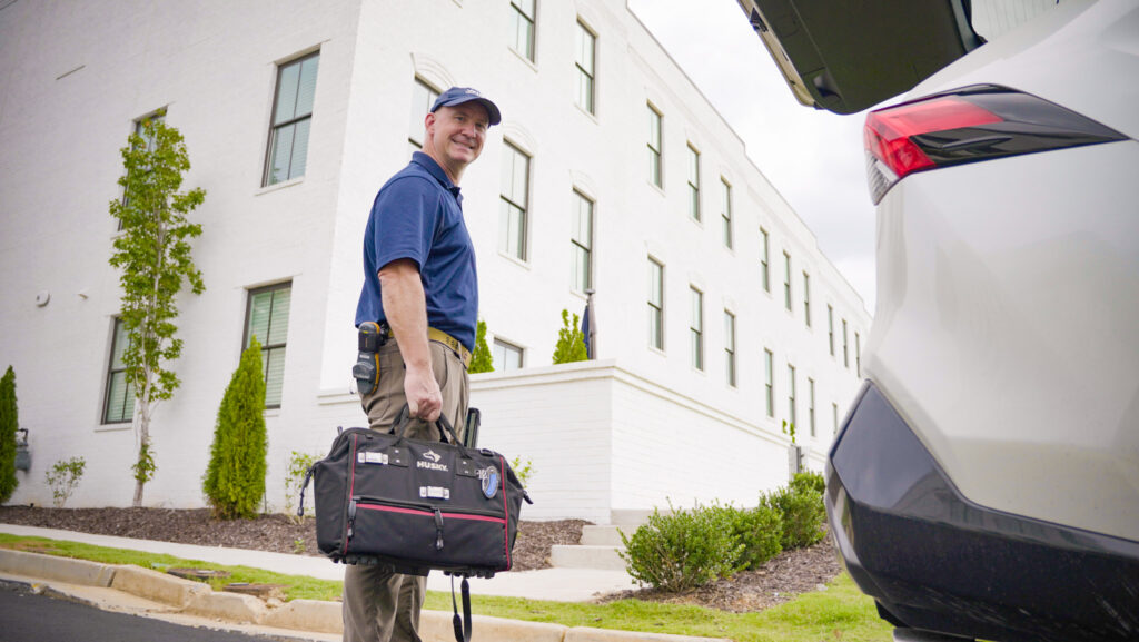 Home Inspector wearing a blue cap and polo carrying a toolbag walks past a white building towards an open car trunk. 