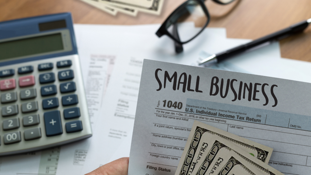 Close-up image of a hand holding a document labeled "Small Business" and a U.S. 1040 tax form. A calculator, glasses, and cash are visible on a desk.