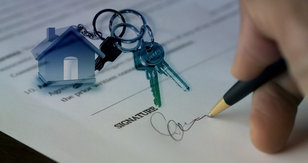 A person signs a document next to a set of house keys and a house-shaped keychain, suggesting a real estate or home purchase agreement.