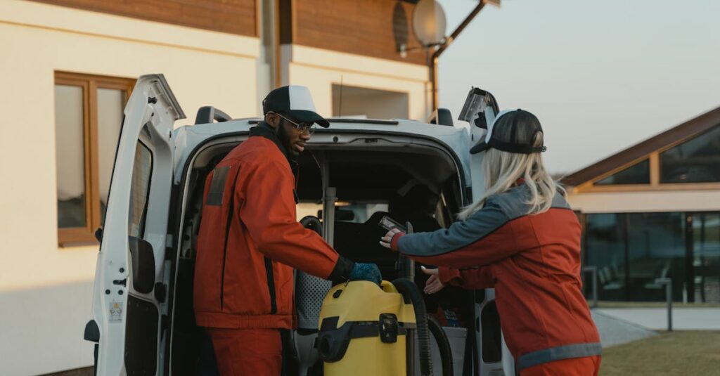 Two cleaners in red uniforms load a vacuum into a white van outdoors.